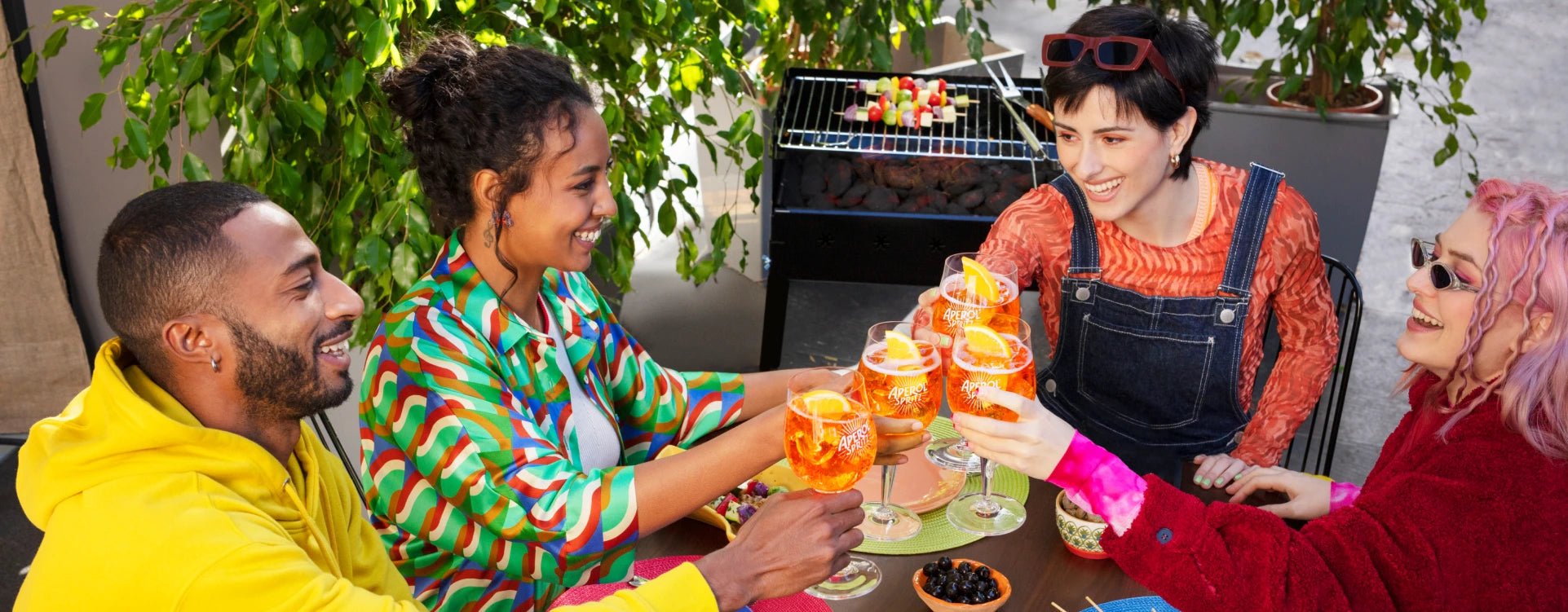 Four friends clinking Aperol Spritz glasses at an outdoor barbecue, colorful outfits and joyful vibe.